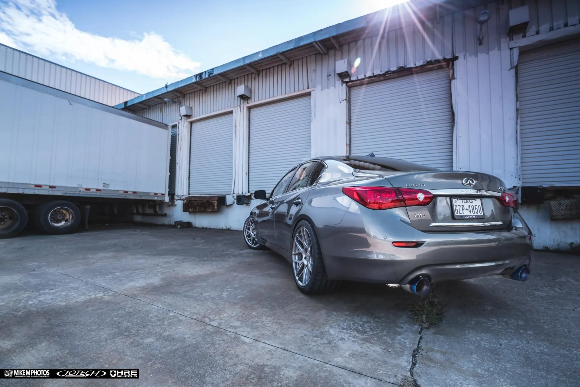 Grey Infiniti Q50 sedan with aftermarket wheels and blue-tipped exhaust parked in front of loading docks.