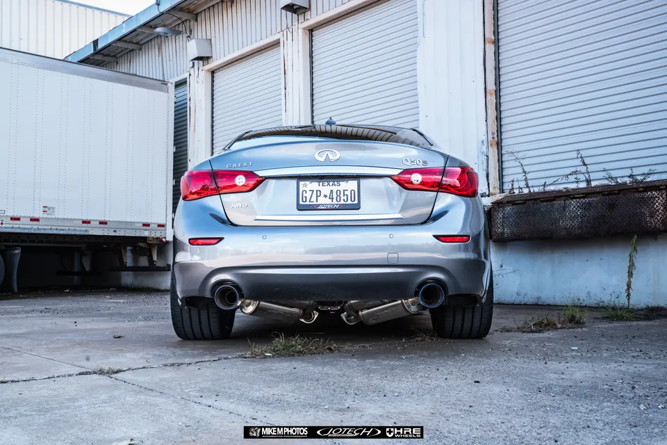 Rear view of a silver Infiniti Q50S with an aftermarket dual exhaust system parked in an industrial area.