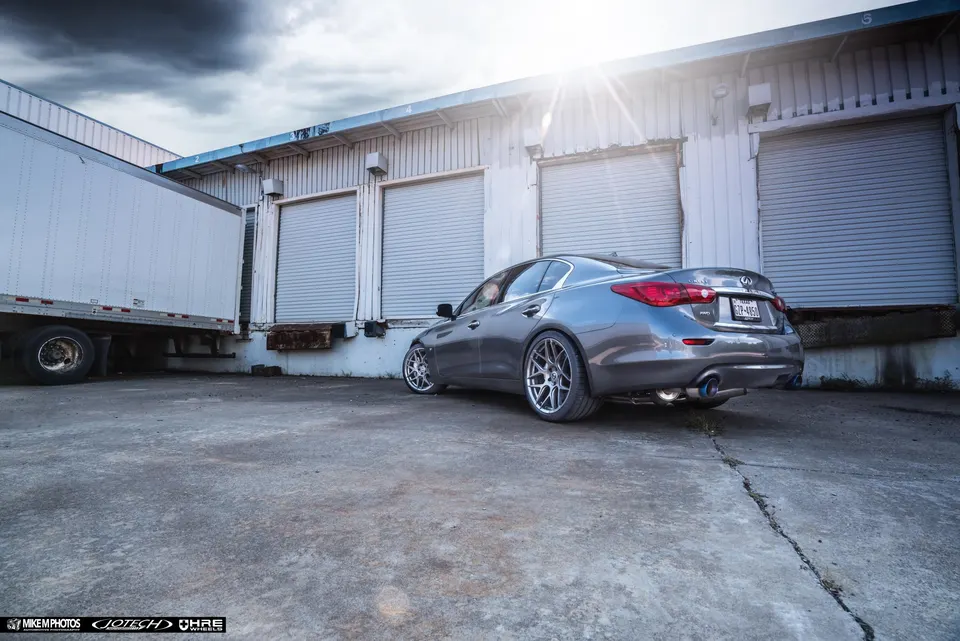 A grey Infiniti Q50 sedan with aftermarket wheels and a performance exhaust system parked in front of a loading dock.