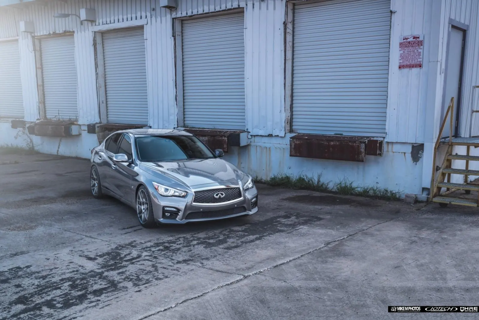 A silver Infiniti Q50 sedan with aftermarket wheels parked in front of a loading dock with multiple garage doors.