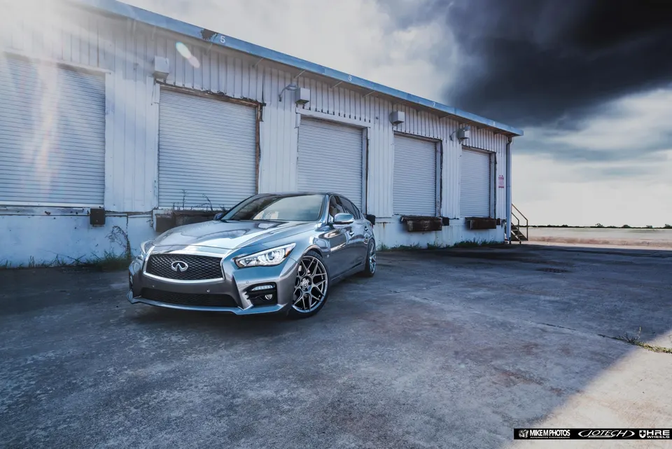 A silver Infiniti Q50 sedan with custom HRE wheels parked in front of a white industrial building under a dramatic sky.