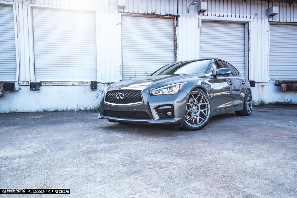 Grey Infiniti Q50 sedan with aftermarket multi-spoke wheels parked in front of a warehouse with roll-up doors.