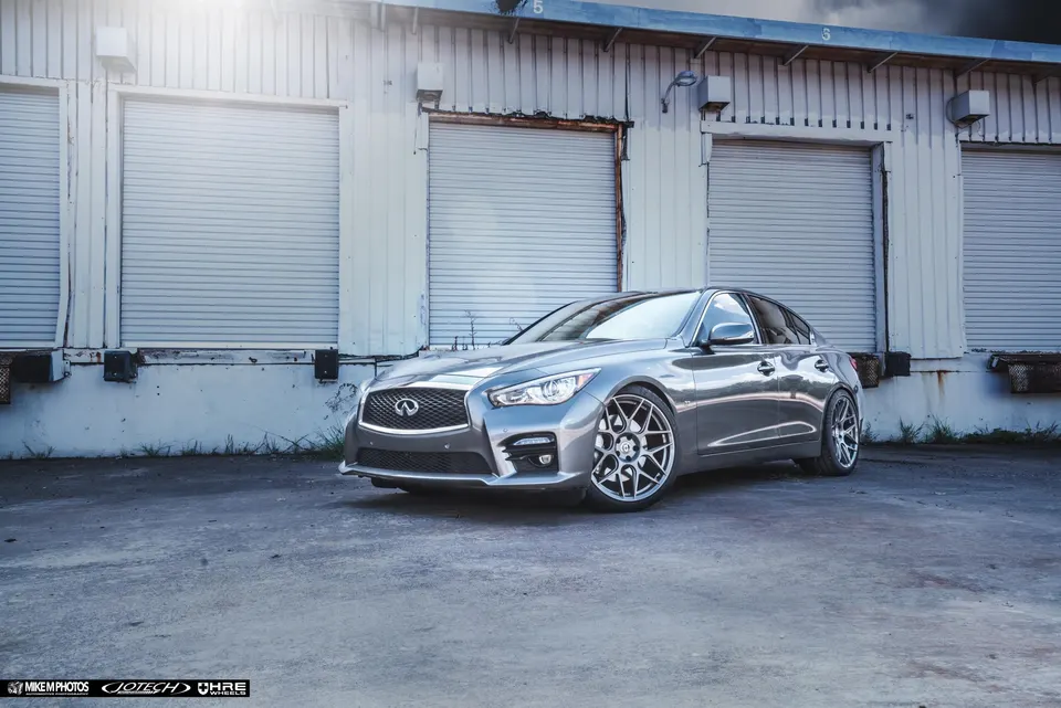 Silver Infiniti Q50 sedan with aftermarket multi-spoke wheels parked in front of a warehouse building.