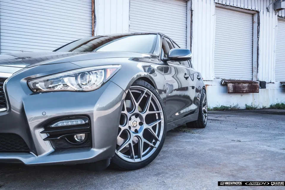 A grey Infiniti Q50 sedan with aftermarket multi-spoke wheels parked in front of a garage door.