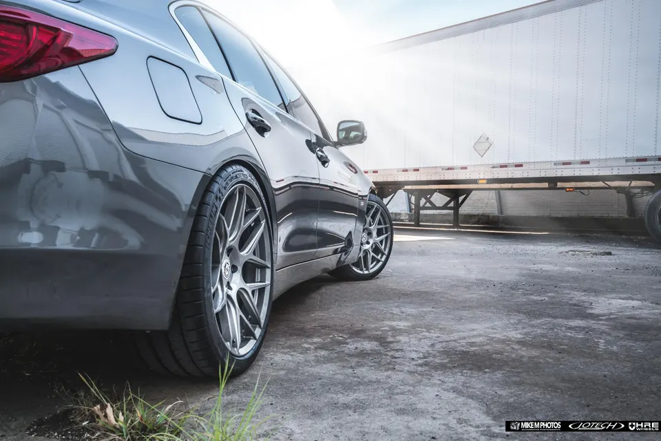 Grey Infiniti Q50 sedan with aftermarket HRE wheels and performance tires parked outdoors next to a semi-trailer.