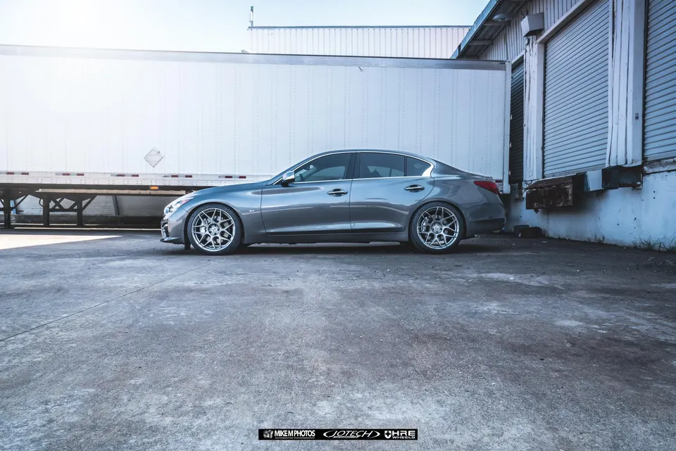 Grey Infiniti Q50 sedan with aftermarket wheels and lowered suspension parked next to a semi-trailer and loading dock.