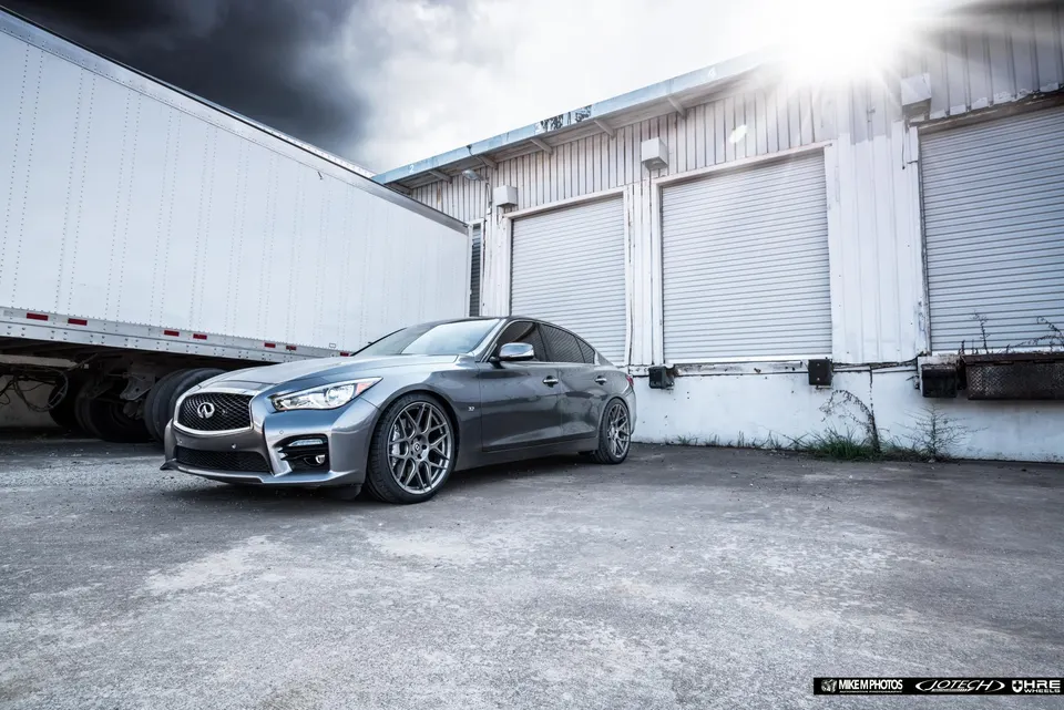 Grey Infiniti Q50 sedan with aftermarket wheels parked in an industrial loading dock area under dramatic skies.
