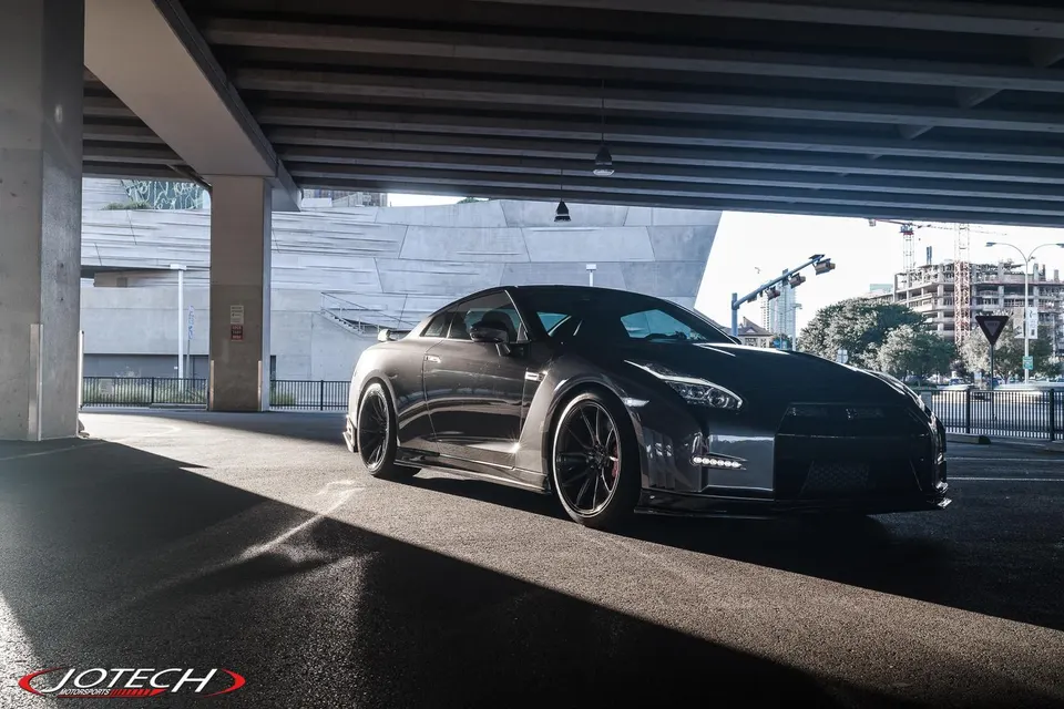 A dark grey Nissan GT-R with aftermarket wheels parked under an overpass, sunlight highlighting its sleek design.