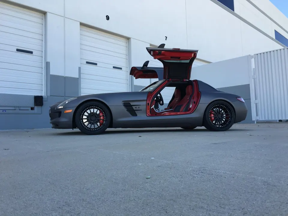 Matte grey Mercedes-Benz SLS AMG with gullwing doors open, custom black wheels, and red interior parked outside a building.