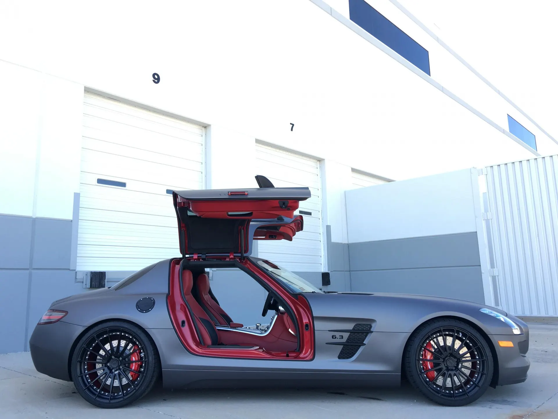 Matte grey Mercedes-Benz SLS AMG with gullwing doors open, showcasing red interior and custom black wheels with red calipers, parked outside a shop.