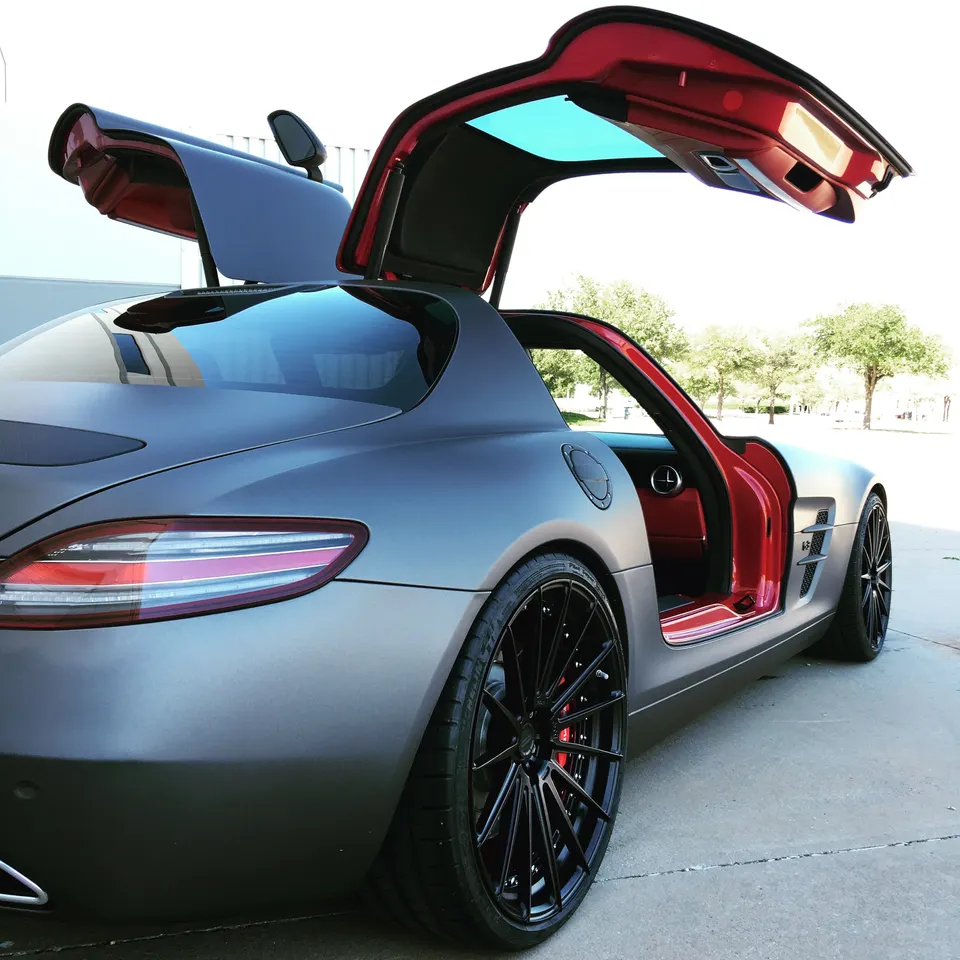 Matte grey Mercedes-Benz SLS AMG with open gullwing doors, custom black wheels, and red brake calipers parked outdoors.