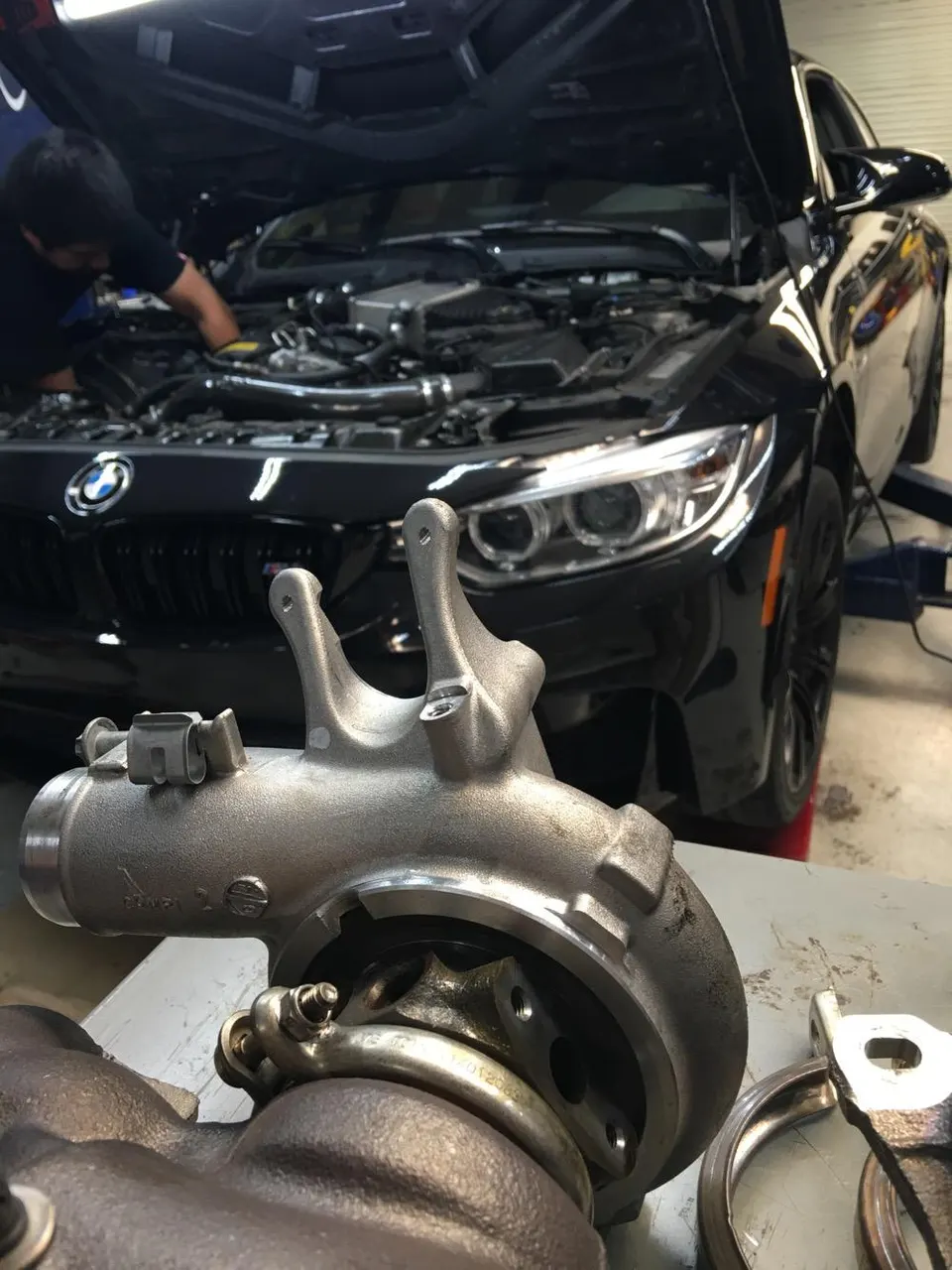 A black BMW M4 with its hood open in a shop, with a mechanic working on the engine and a turbocharger in the foreground.