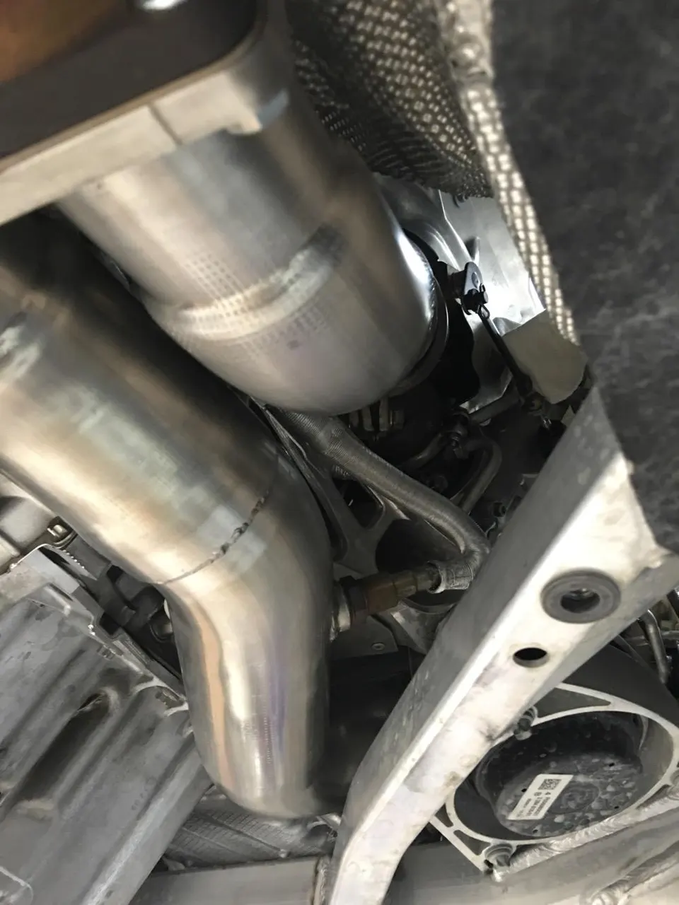 Close-up of a stainless steel exhaust downpipe and underbody components of a performance car.