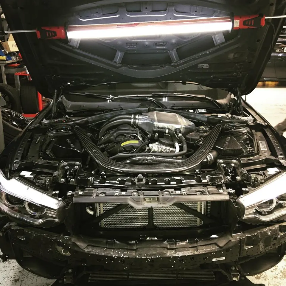 BMW M series car in a shop with the hood open and front bumper removed, showcasing the engine bay, intercooler, and carbon fiber brace.