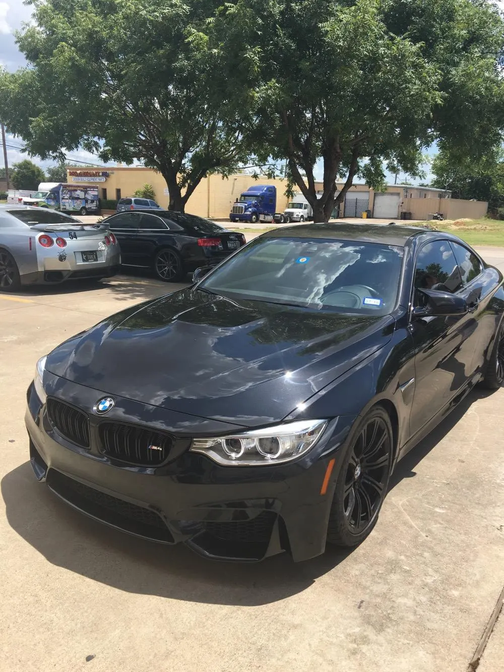 A black BMW M4 coupe parked in a sunny parking lot with a silver Nissan GT-R in the background.
