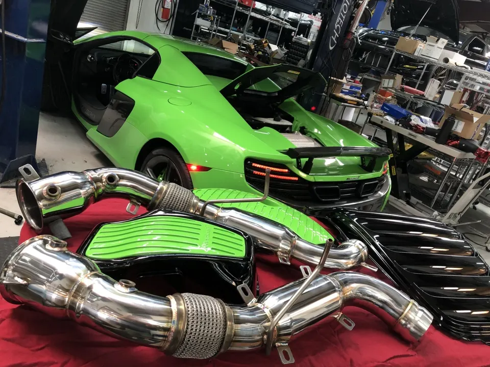 A bright green McLaren supercar in a performance tuning shop with aftermarket exhaust downpipes displayed in the foreground.