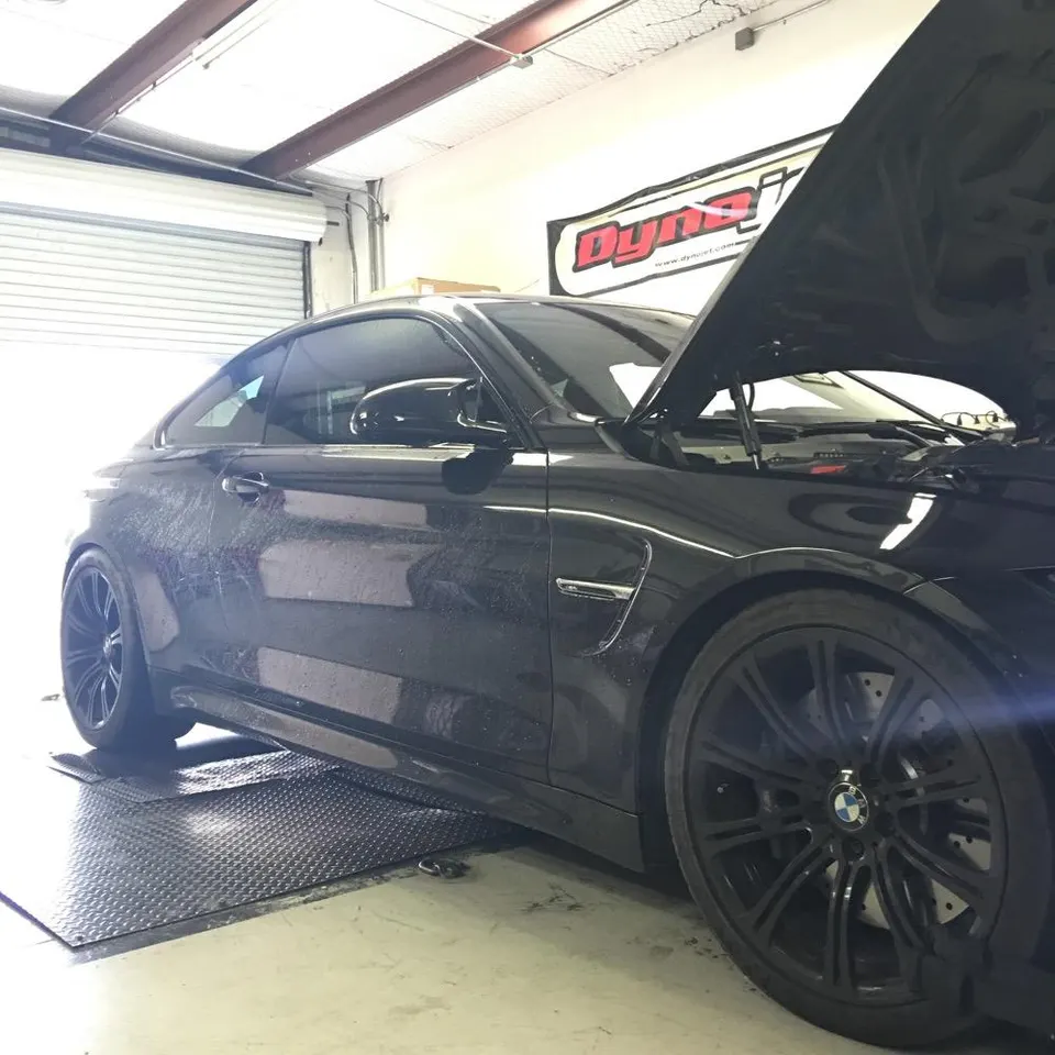 Black BMW M4 on a Dynojet dynamometer with the hood open, undergoing performance testing at a tuning shop.