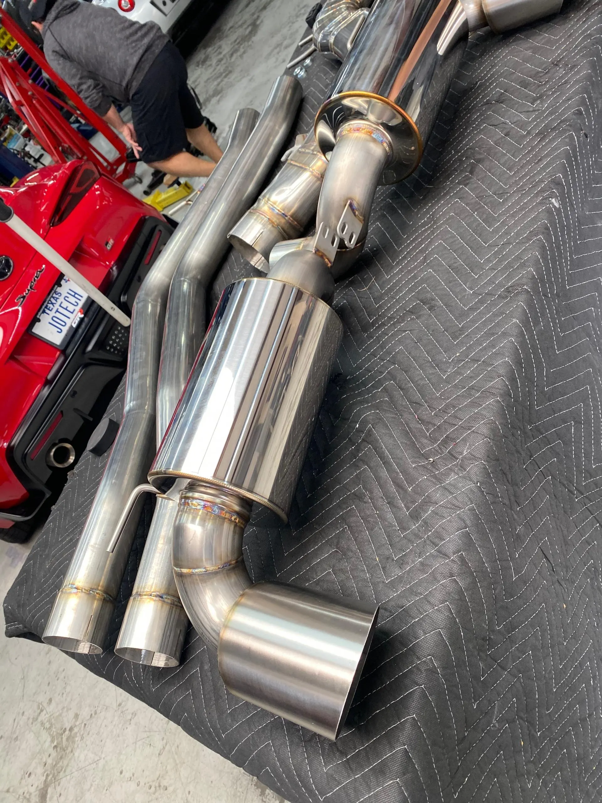 Stainless steel exhaust system components laid out on a blanket in a performance shop, with a red Toyota Supra in the background.