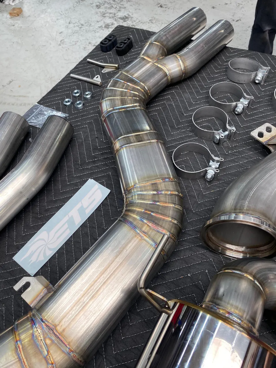 ETS stainless steel exhaust system components laid out in a shop, showing pipes, muffler, and clamps ready for installation.
