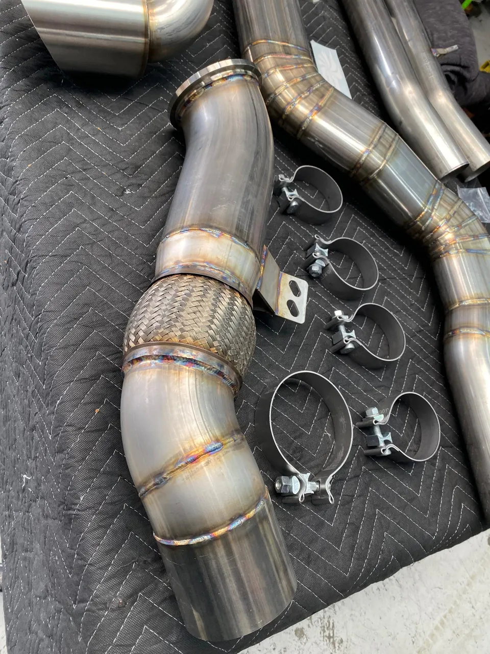 Stainless steel exhaust pipes with visible welds and v-band clamps laid on a black mat, ready for installation.