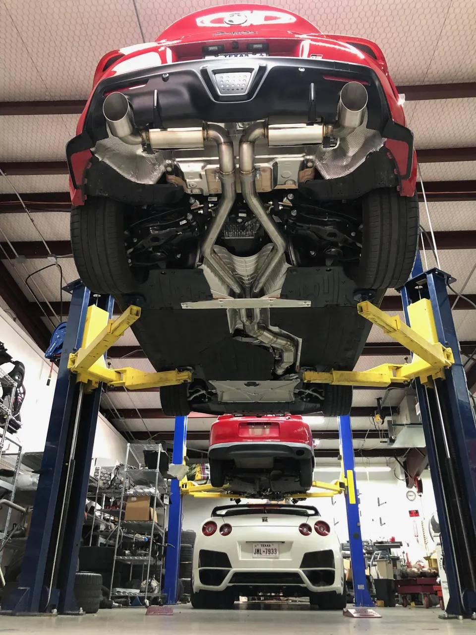 A red Toyota Supra on a car lift showcasing its aftermarket exhaust system, with a white Nissan GT-R below in a performance shop.