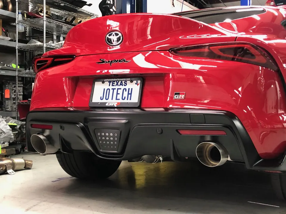 Red Toyota Supra GR with aftermarket exhaust tips and a Texas Jotech license plate in a performance shop.