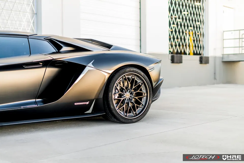 Rear side view of a matte black Lamborghini Aventador with custom HRE wheels parked outside an industrial building.