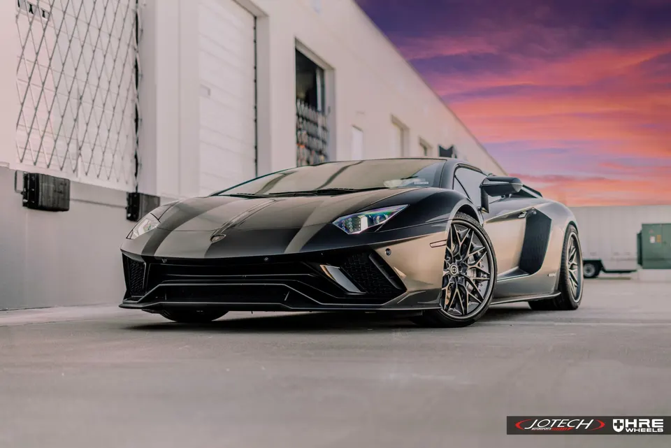 A black Lamborghini Aventador with custom wheels parked in front of a building at sunset.