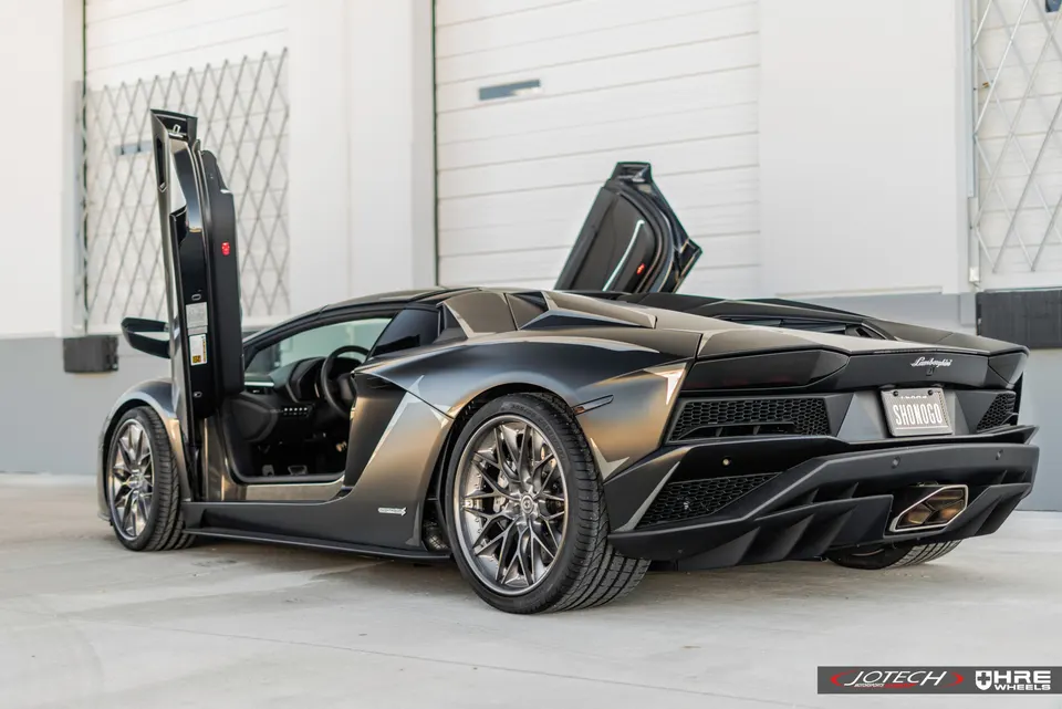 Dark grey Lamborghini Aventador with open scissor doors and aftermarket HRE wheels in a clean setting.