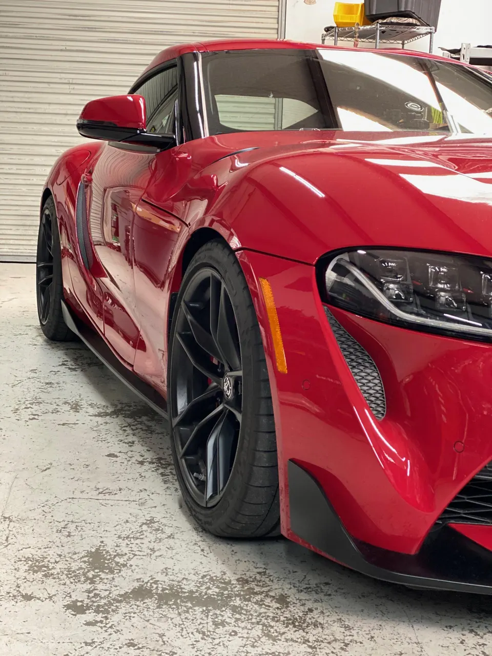 A low-angle side profile of a red Toyota Supra with custom black wheels and a lowered stance in a garage.