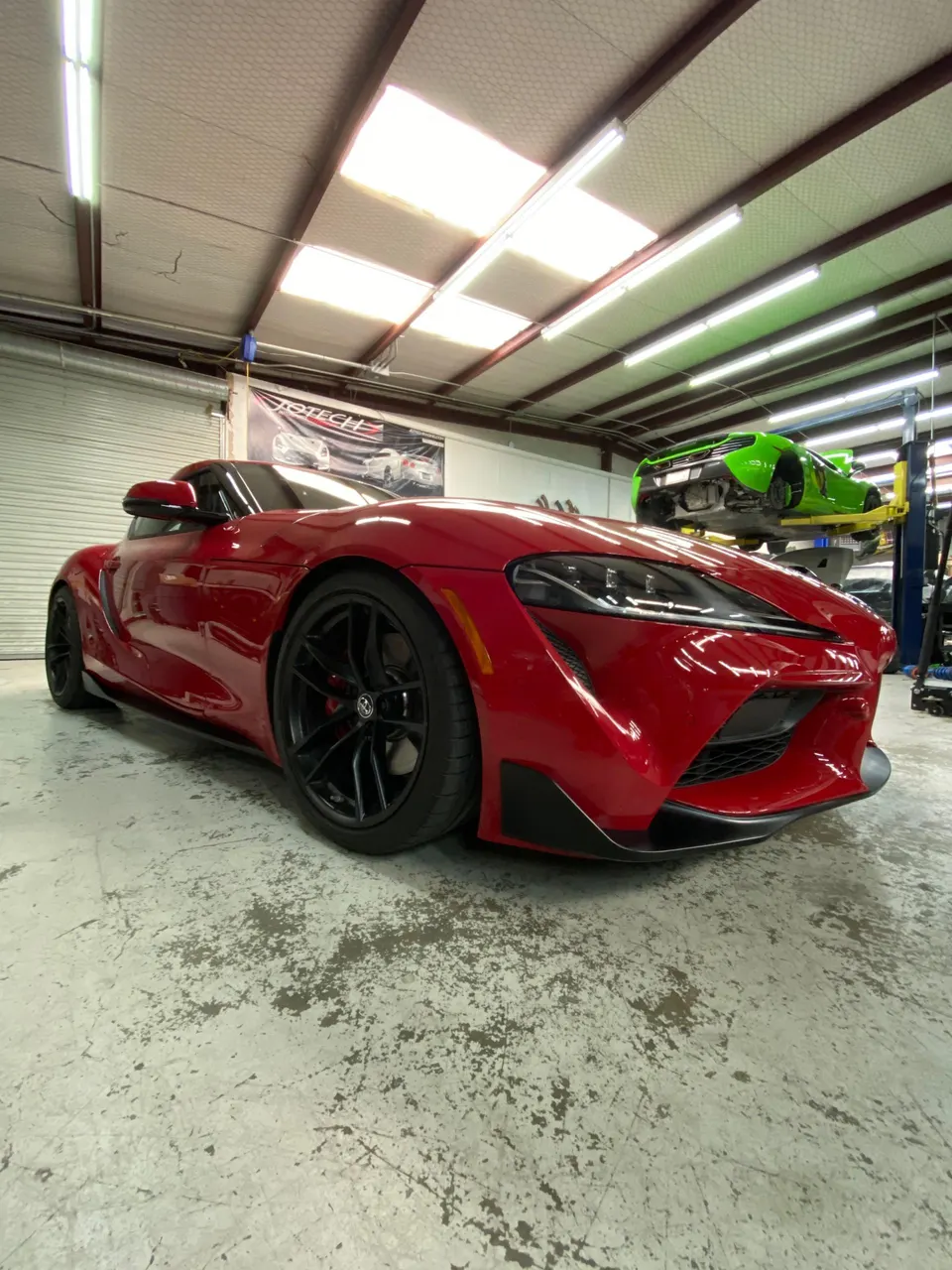 Red Toyota Supra parked in a Jotech Motorsports performance tuning shop with a green McLaren on a lift in the background.