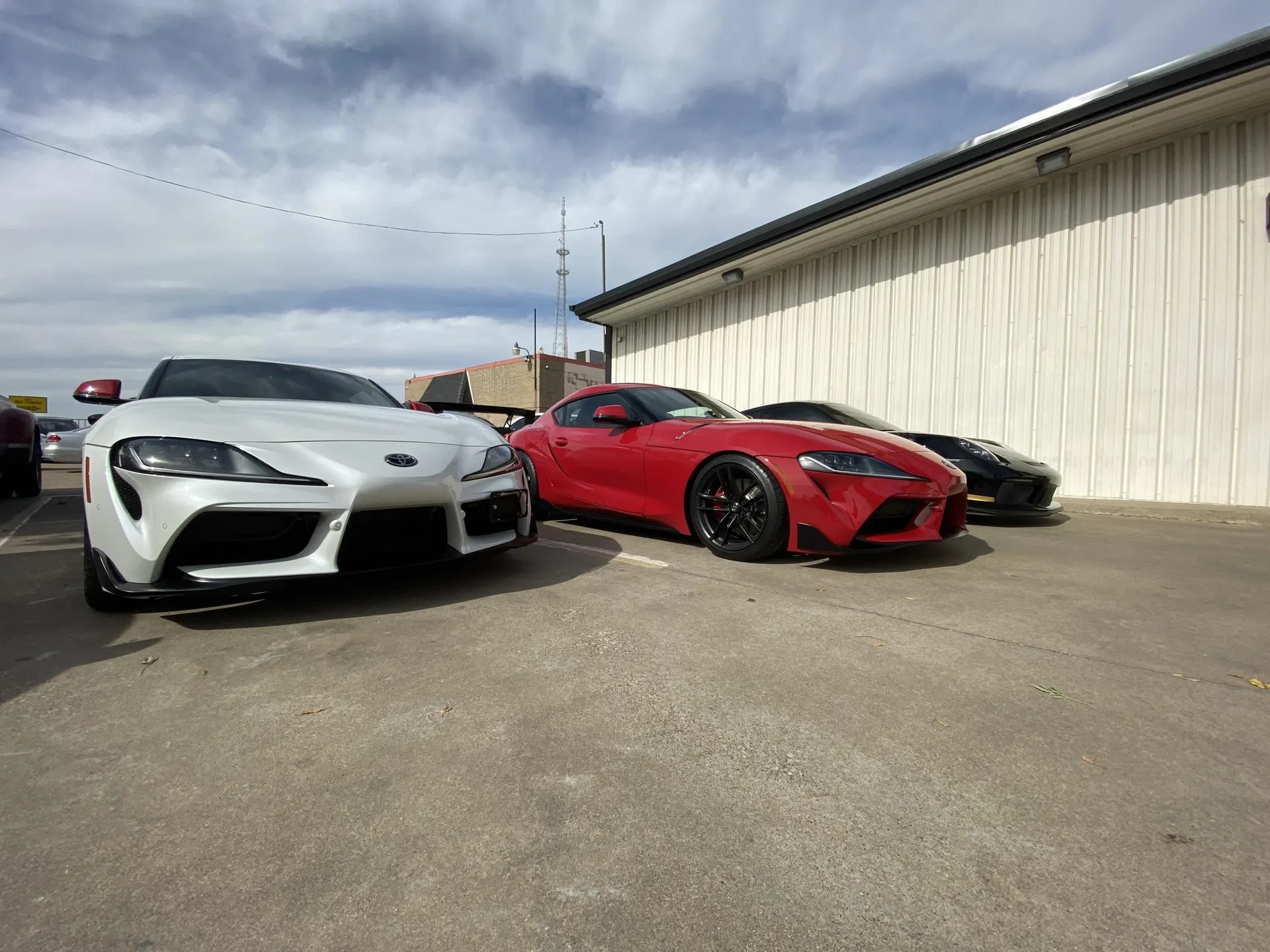 White and red Toyota Supra sports cars parked outdoors in a lot, with a black sports car partially visible.