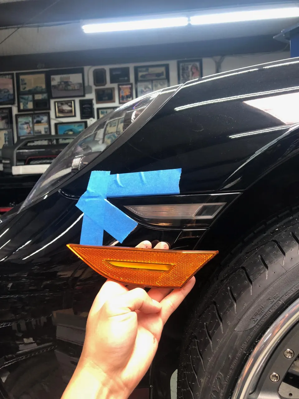 A hand holding an amber side marker light next to the fender of a black car, with blue painter's tape on the body.