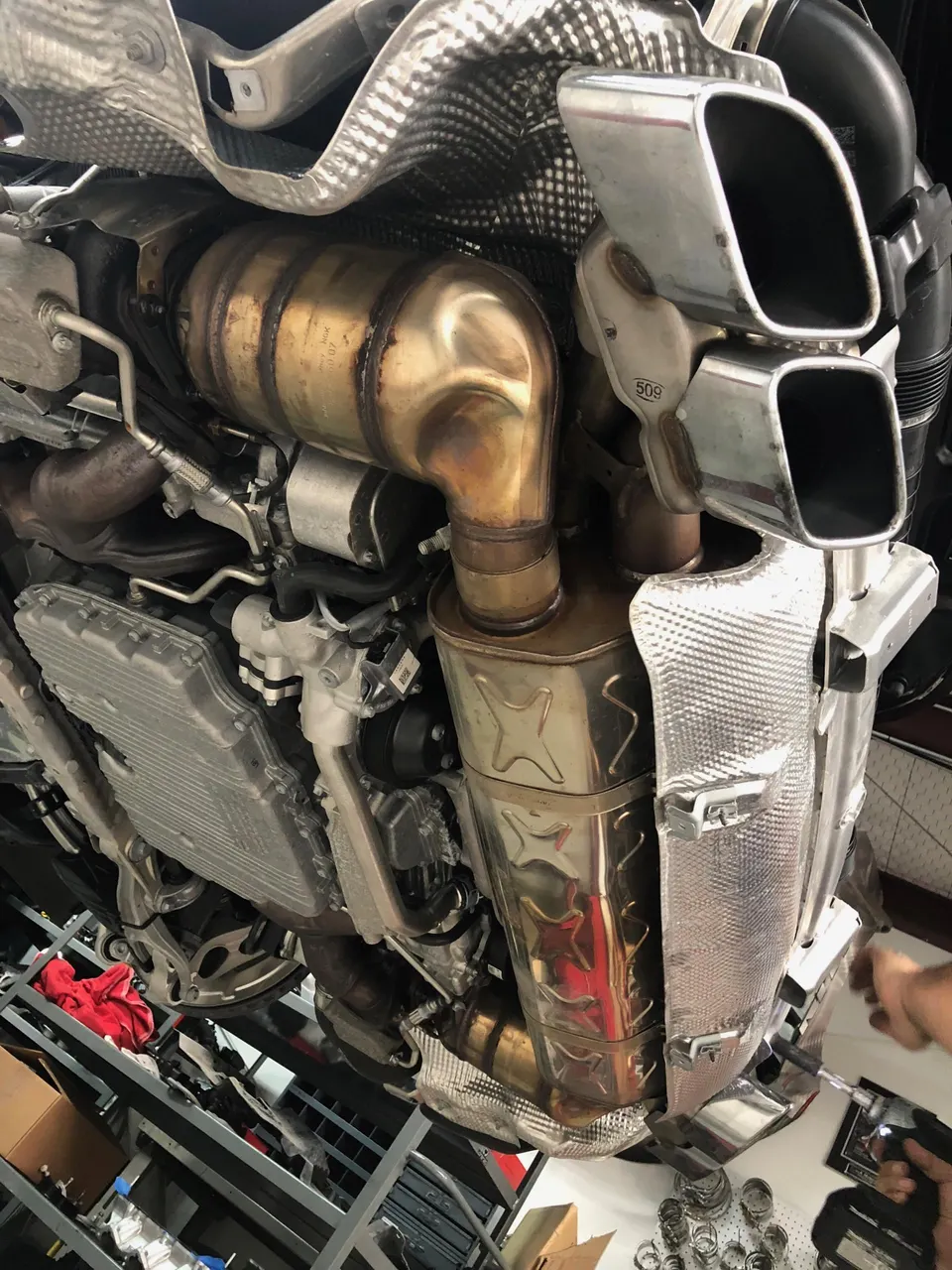 Underside view of a car on a lift, showcasing the exhaust system with catalytic converter, muffler, and quad exhaust tips.