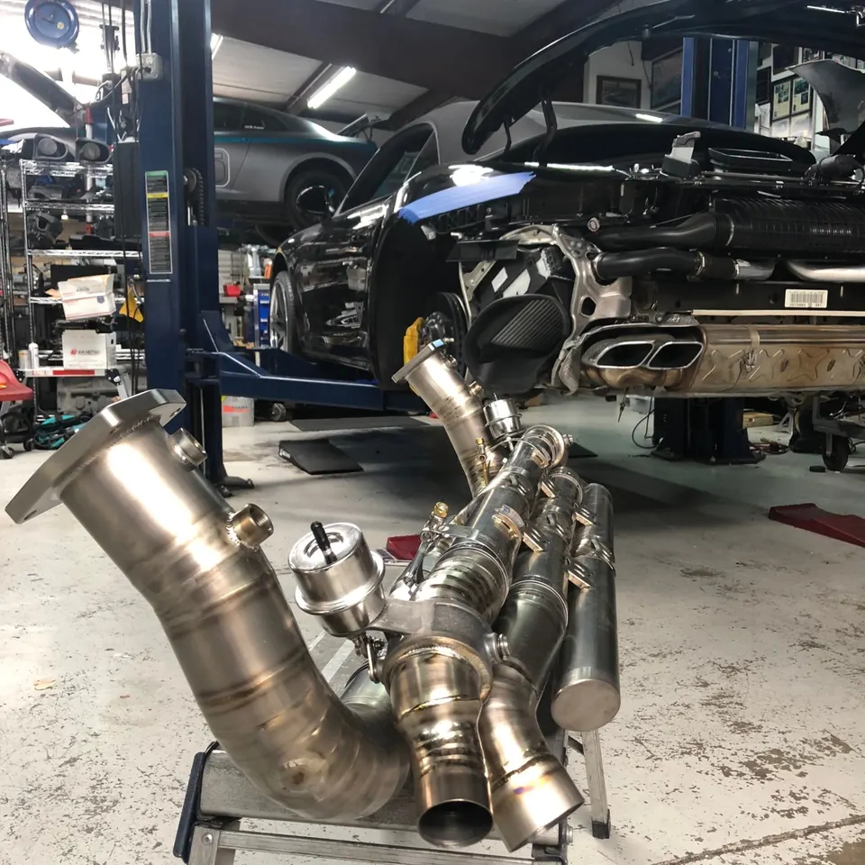 A Porsche 911 on a lift in a performance shop with a high-performance exhaust system on a stand in the foreground.