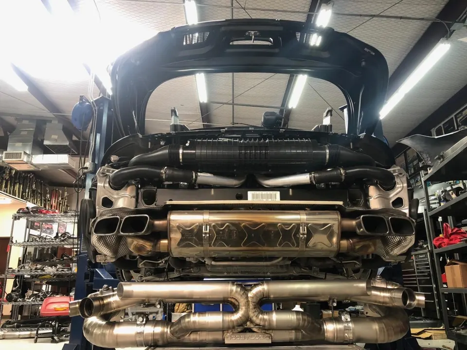 Underside view of a Porsche 911 on a lift in a performance shop, showcasing the stock and aftermarket exhaust systems.