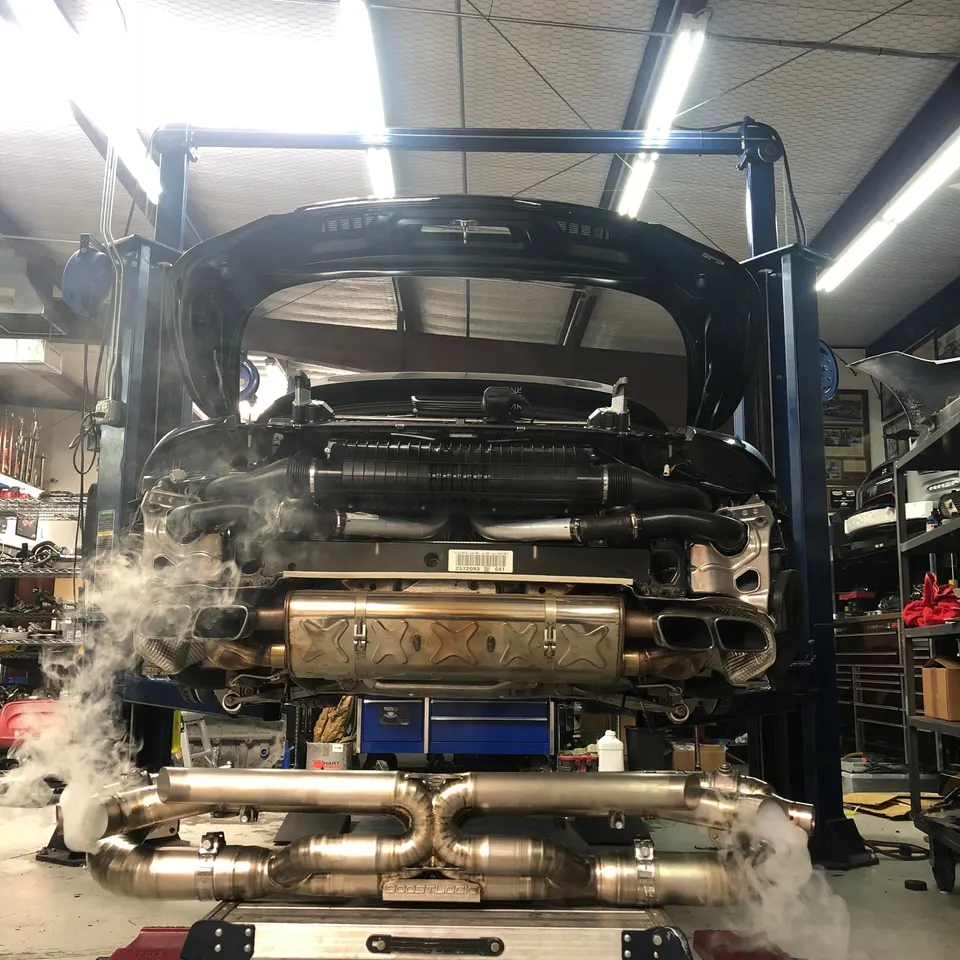 A black Porsche on a lift in a shop with its exhaust system being worked on, showing a new exhaust on the floor with smoke.