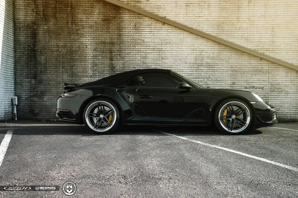 Black Porsche 911 convertible with HRE wheels and yellow brake calipers parked against a brick wall.