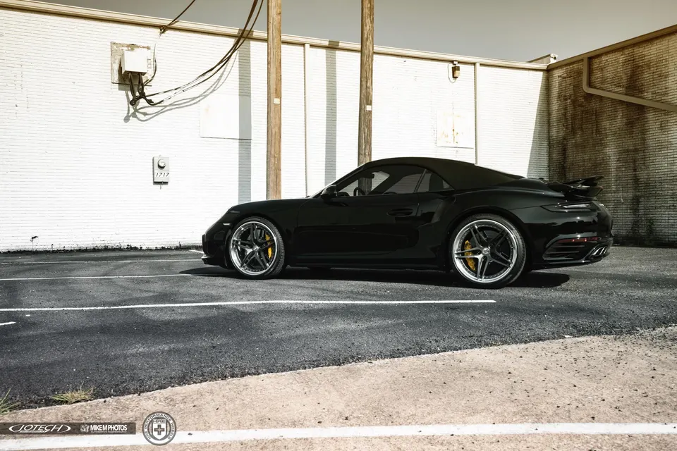 A black Porsche 911 convertible with custom wheels and yellow brake calipers parked next to a white brick wall.