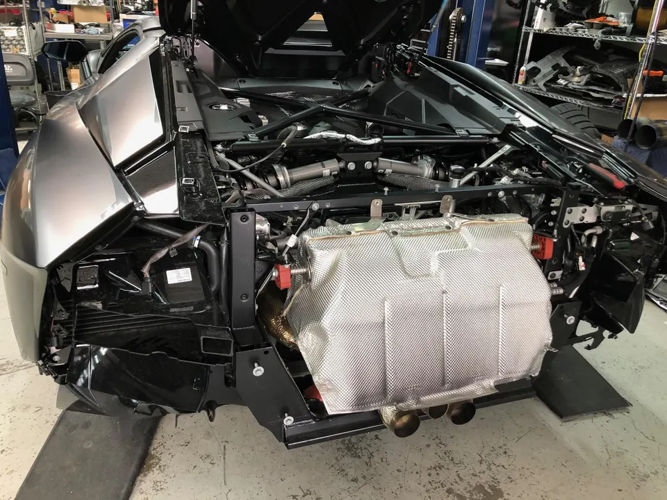 Rear view of a Lamborghini Aventador in a shop with the bumper removed, exposing the engine and exhaust system.