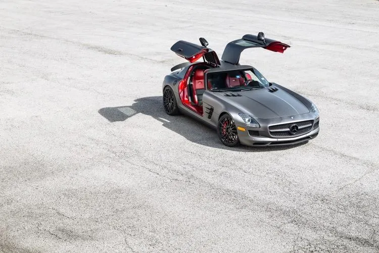 Grey Mercedes-Benz SLS AMG with gullwing doors open, showcasing custom wheels on a clean concrete surface.