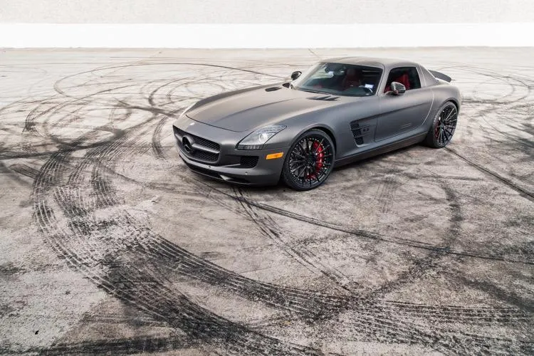 Matte grey Mercedes-Benz SLS AMG with custom black wheels and red brake calipers parked on a concrete lot with tire marks.