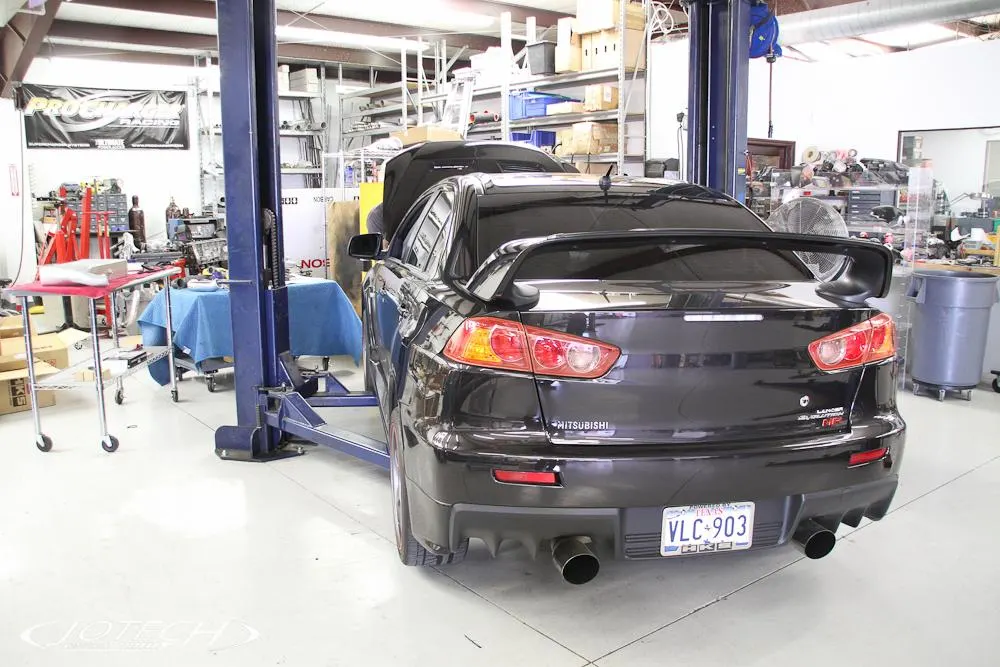 A black Mitsubishi Lancer Evolution X on a car lift in a Jotech Motorsports shop, showcasing its rear exhaust and diffuser.