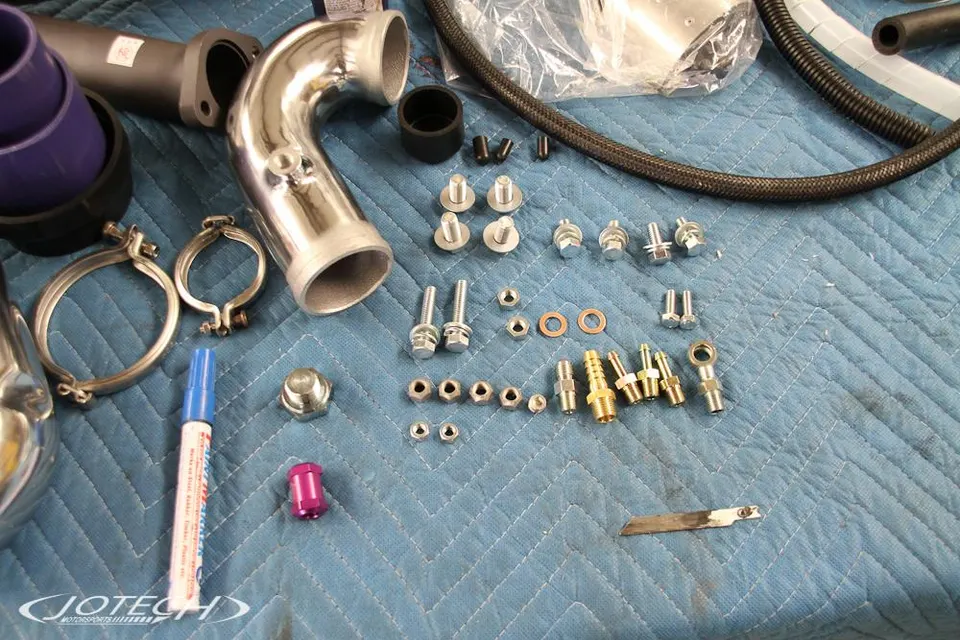 Various performance car parts including polished boost piping, V-band clamps, and fittings laid out on a blue mat at Jotech Motorsports.