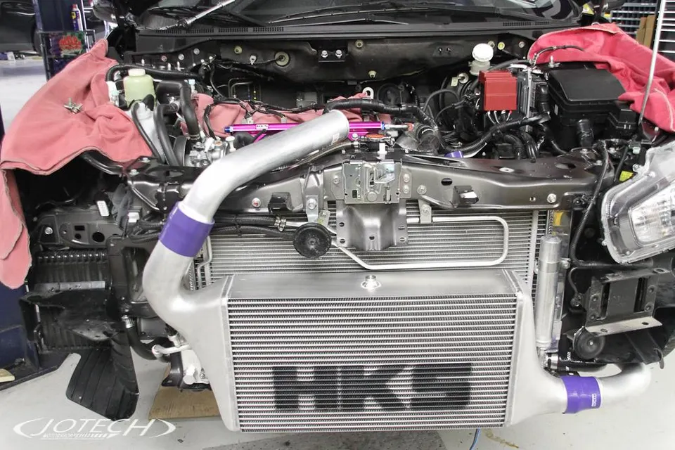 Front view of a car in a shop with the bumper removed, showcasing a large HKS front mount intercooler and custom boost piping installation.