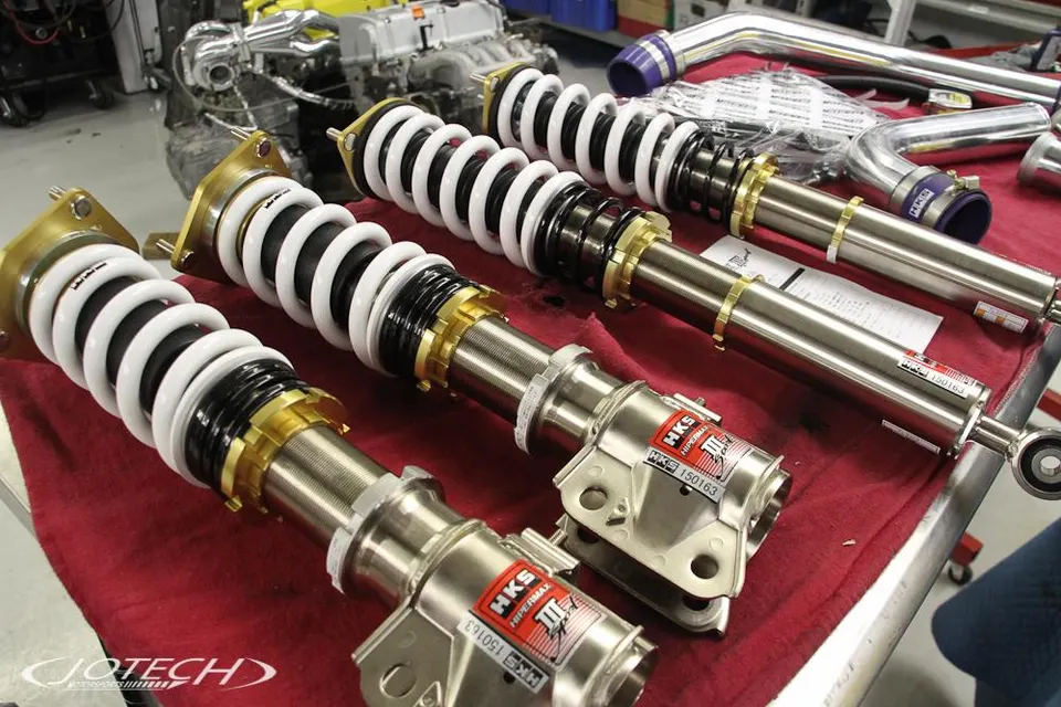 HKS Hipermax coilovers and performance piping laid out on a workbench in a Jotech Motorsports shop.