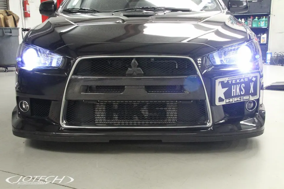 A black Mitsubishi Lancer Evolution X with bright headlights, a carbon fiber front grille, and a large aftermarket intercooler.