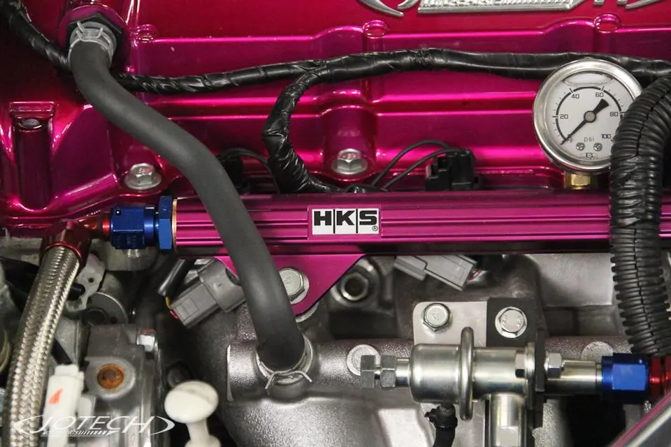 A close-up of a pink HKS fuel rail, fuel pressure gauge, and fuel lines in a performance engine bay.