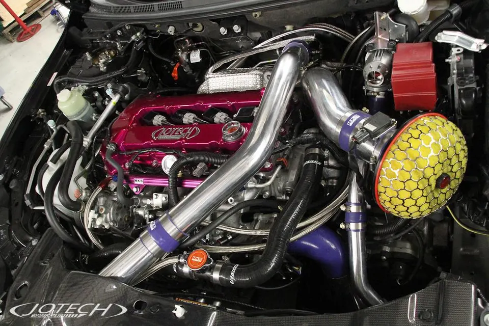 A detailed view of a Jotech Motorsports modified engine bay featuring a pink valve cover, HKS intake, and polished boost piping.