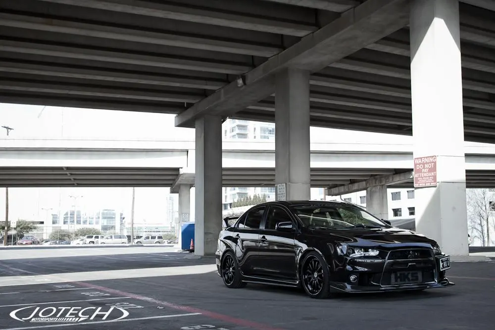 Black Mitsubishi Lancer Evolution X with aftermarket wheels and HKS front plate parked under a concrete overpass.